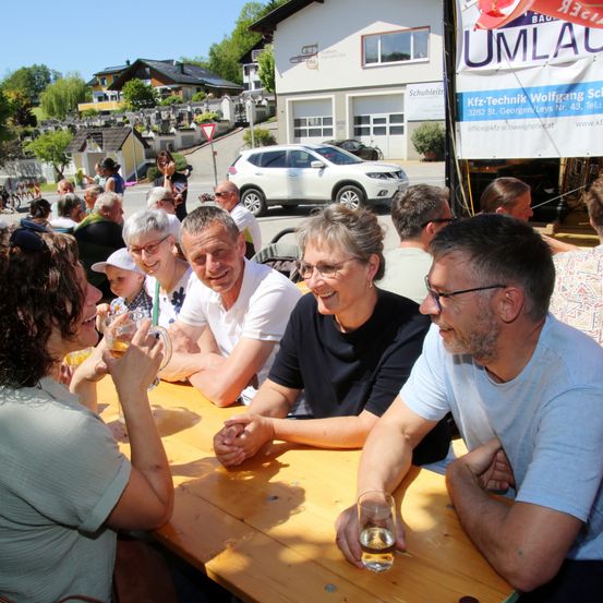 Eine Gruppe von Erwachsenen, einige mit Brille, sitzt um einen Tisch. Ein Mann in einem blauen T-Shirt lächelt und hält ein Glas. Eine Frau in einem schwarzen T-Shirt schaut ihn an. Ein Banner mit dem Text "UMLAU" ist im Hintergrund.