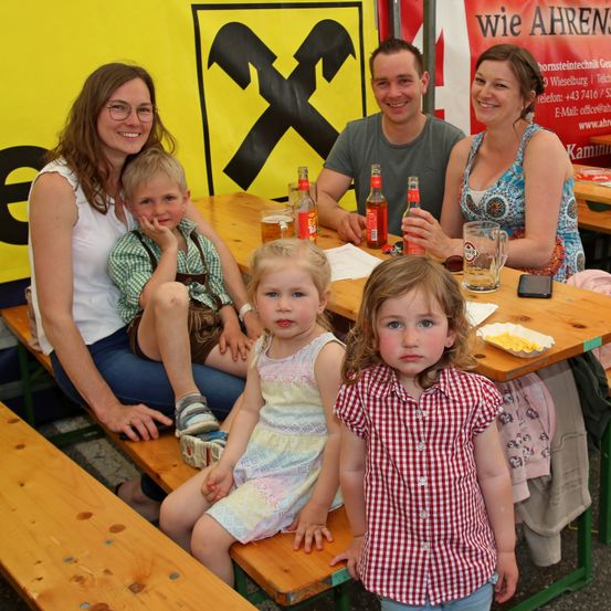 Eine Familie mit fünf Personen sitzt an einem Picknicktisch im Freien. Sie lächeln für ein Foto. Der Mann und die Frau tragen Freizeitkleidung. Vor ihnen sitzen zwei kleine Kinder. Flaschen Bier stehen auf dem Tisch.