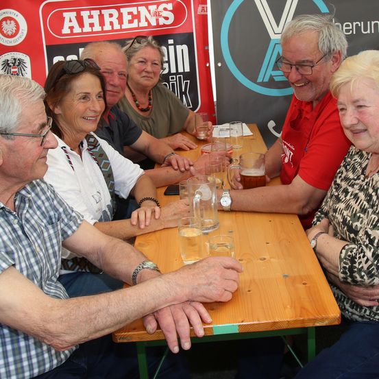 Eine Gruppe von Menschen sitzt um einen Tisch mit Getränken und lächelt, in der Nähe eines Banners mit dem Text 'Ahrens'. Sie tragen Freizeitkleidung und Brillen. Ein Logo und eine Telefonnummer sind auf dem Banner.