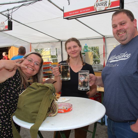 Drei Menschen stehen unter einem Zelt und lächeln. Zwei Frauen halten Biergläser, eine hat eine grüne Tasche. Ein Mann trägt ein blaues Hemd mit weißem Text. Ein Kaiser-Schild ist über ihnen.