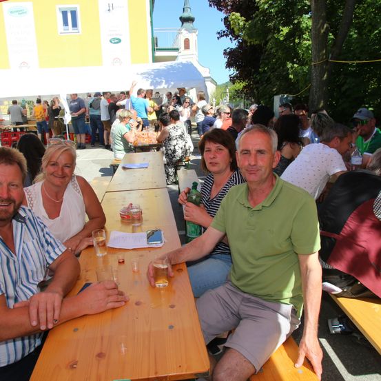 Eine Gruppe von Menschen sitzt an einem langen Tisch im Freien, lächelt und hält Getränke in den Händen. Dahinter versammelt sich eine Menschenmenge, einige stehen, andere sitzen. Im Hintergrund ist ein Gebäude mit einem Turm und einem Zelt zu sehen.