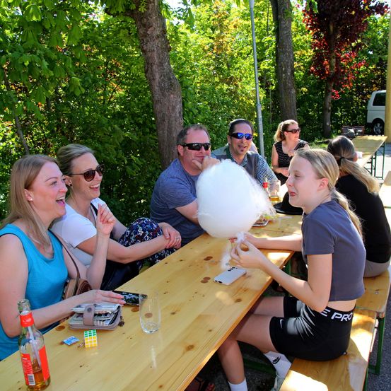 Eine Gruppe Erwachsener macht draußen ein Picknick. Eine Person bläst eine Zuckerwatte auf. Sie sitzen auf Holzbänken unter einem Baum.