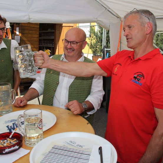 Zwei Männer stehen unter einem Zelt, einer hält ein großes Bierglas. Sie lächeln und scheinen für ein Foto zu posieren. Ein Tisch vor ihnen hat Teller, ein Glas und eine Zigarettenasche.