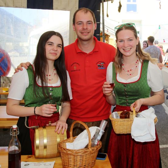 Zwei Frauen in traditioneller deutscher Kleidung mit einem Mann, halten Körbe und Gläser und lächeln in einer Außenumgebung.