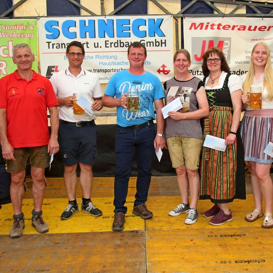 Fünf Personen stehen auf einer Bühne und halten Bierkrüge und Urkunden. Drei Männer und zwei Frauen lächeln. Hinter ihnen sind Banner mit Texten und Logos.