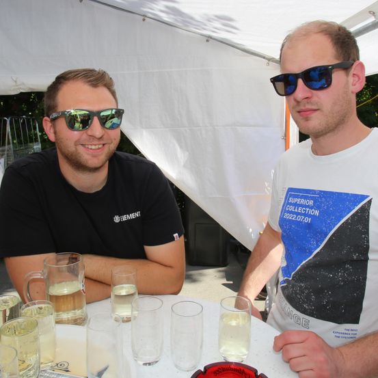 Zwei Männer mit Sonnenbrillen sitzen unter einem weißen Zelt an einem Tisch. Einer trägt ein schwarzes Shirt mit Logo, der andere ein weißes Shirt mit Design. Sie sind von Gläsern mit Getränken umgeben.