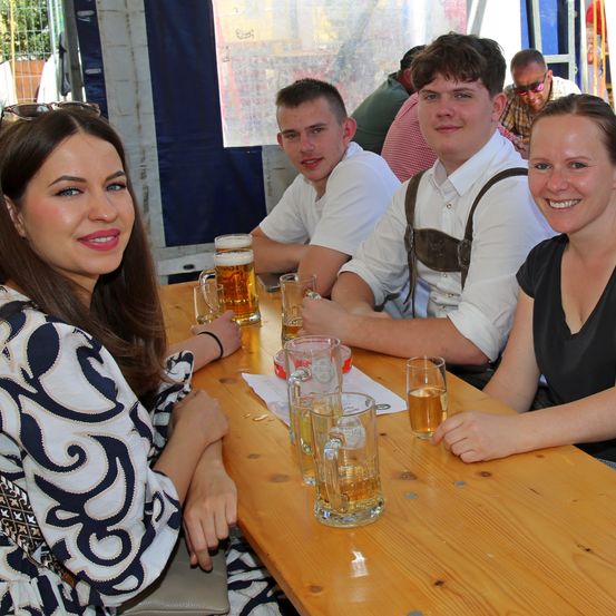 Eine Gruppe von Menschen, darunter zwei Frauen und zwei Männer, sitzen an einem Holztisch und lächeln. Sie haben eine gute Zeit. Auf dem Tisch stehen Biergläser, Gläser und eine Flasche. Hinter ihnen sitzen und stehen weitere Personen. Es gibt ein Zelt mit einer blauen Plane.