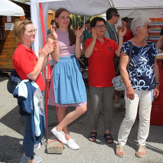 Vier Frauen stehen unter einem Zelt bei einer Veranstaltung im Freien. Sie klatschen, wobei eine Frau einen blauen Rock trägt und die anderen rote Hemden. Hinter ihnen stehen Leute und halten Getränke.