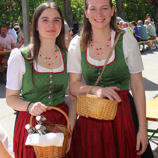 Zwei junge Frauen in traditioneller deutscher Kleidung halten Körbe, lächeln und posieren für ein Foto. Sie tragen grüne und rote Kleider mit Halsketten und Armbändern. Im Hintergrund sitzen und stehen Leute an Tischen.