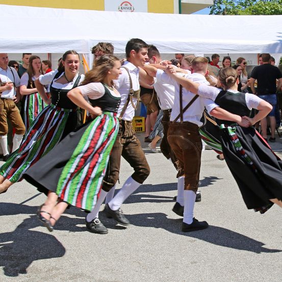 Mehrere Menschen in traditioneller Kleidung tanzen in einem Kreis unter einem weißen Zelt. Zuschauer stehen hinter ihnen, und einige lächeln. Die Schatten der Menschen werden auf den Boden geworfen.