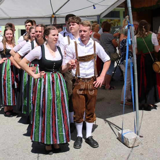 Eine Gruppe junger Leute in traditioneller bayerischer Kleidung steht draußen. Ein Mann in Lederhosen lächelt und interagiert mit einer Frau in einem grünen und weißen Kleid.