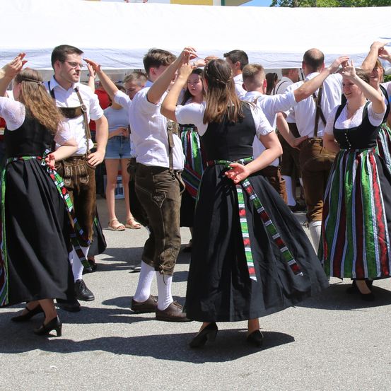 Eine Gruppe von Menschen in traditioneller Kleidung tanzt im Freien unter einem Zelt. Sie lächeln und scheinen Spaß zu haben.