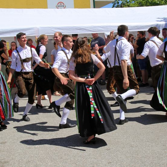 Eine Gruppe von Menschen in traditioneller bayerischer Kleidung tanzt im Freien. Ein großes weißes Zelt steht hinter ihnen, und mehrere Zuschauer beobachten.