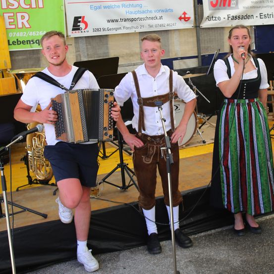 Drei Personen stehen auf der Bühne und spielen mit Musikinstrumenten. Der Mann auf der linken Seite spielt Akkordeon, der Mann in der Mitte trägt traditionelle deutsche Kleidung und die Frau auf der rechten Seite singt in ein Mikrofon.