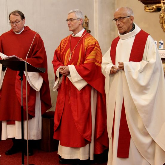 Drei Priester in roten und weißen Gewändern stehen in einer Kirche, einer liest aus einem Buch an einem Pult.