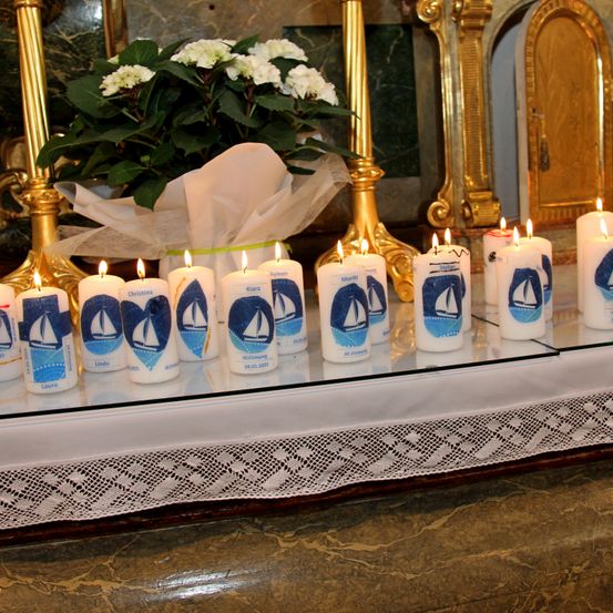 Eine Reihe personalisierter Kerzen mit Segelboot-Designs und Namen wie Laura, Linda und anderen sind auf einem Glasregal vor einem goldenen Altar angeordnet. Hinter den Kerzen steht ein weißer Blumenstrauß.