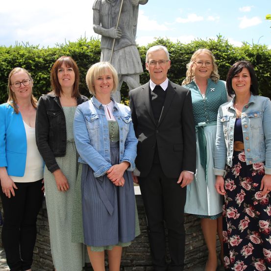 Eine Gruppe von Menschen steht vor einer Statue eines Soldaten. Sie alle lächeln und scheinen für ein Foto zu posieren. Dahinter stehen Büsche.