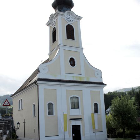 Eine kleine, traditionelle Kirche mit Glockenturm steht gegen einen bewölkten Himmel. Das Gebäude hat ein beiges Äußeres mit weißen Akzenten und verfügt über Rundbogenfenster und eine schwarze Tür.
