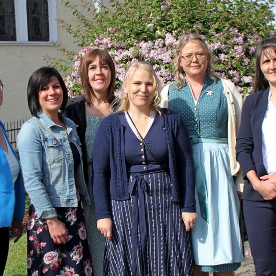 Eine Gruppe von Frauen steht lächelnd draußen. Sie stehen vor einem Gebäude mit weißer Wand und rosa Blumen. Sie tragen verschiedene Outfits, von lässig bis geschäftlich.