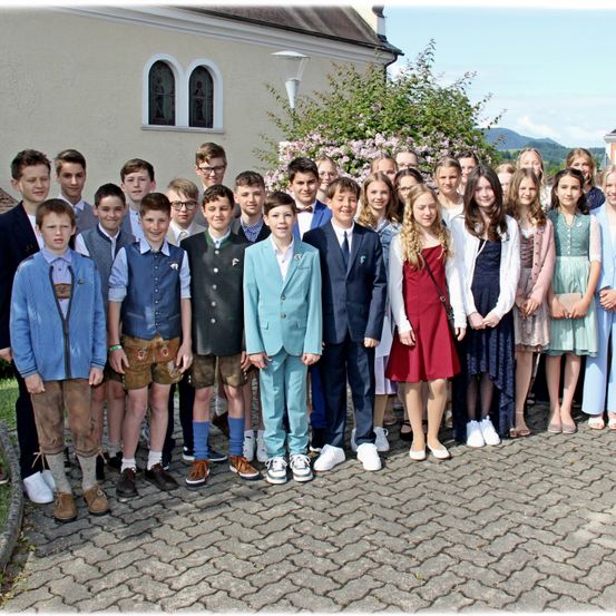 Eine Gruppe von Schülern in traditioneller Kleidung steht vor einem Gebäude mit Bäumen und Bergen im Hintergrund.