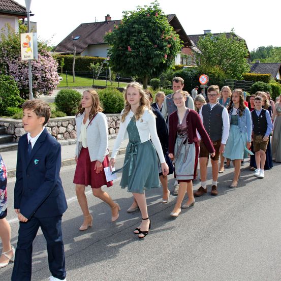 Eine Gruppe von Menschen, überwiegend junge Mädchen, geht in einer Reihe auf einer Straße. Sie tragen traditionelle Kleidung, einige in Röcken und hohen Schuhen. Im Hintergrund befinden sich Häuser und Grünflächen.