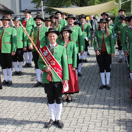 Eine Musikkapelle in grünen Uniformen steht im Freien. Ein Mann und eine Frau vorne halten eine rote Schärpe. Dahinter spielt ein Mann eine Posaune. Bäume und ein Zelt sind im Hintergrund.