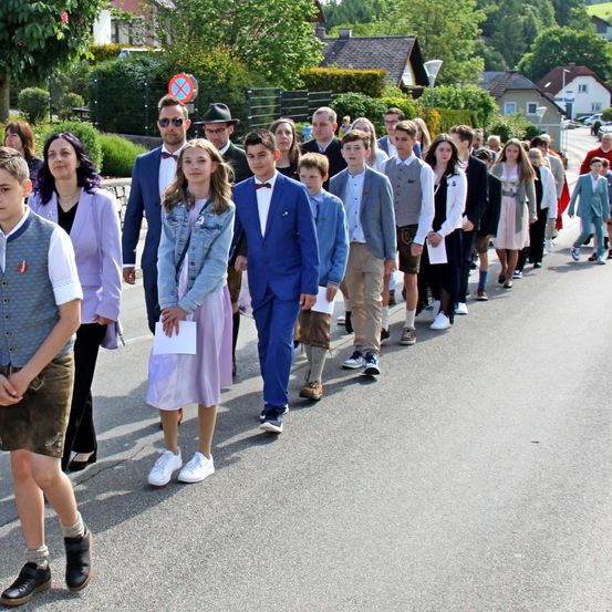 Eine Gruppe von Personen, einige Erwachsene und einige Kinder, gehen in einer Reihe auf einer Straße. Sie sind in verschiedenen Kleidungsstücken gekleidet und einige halten Papiere. Hinter ihnen stehen Häuser und Bäume.