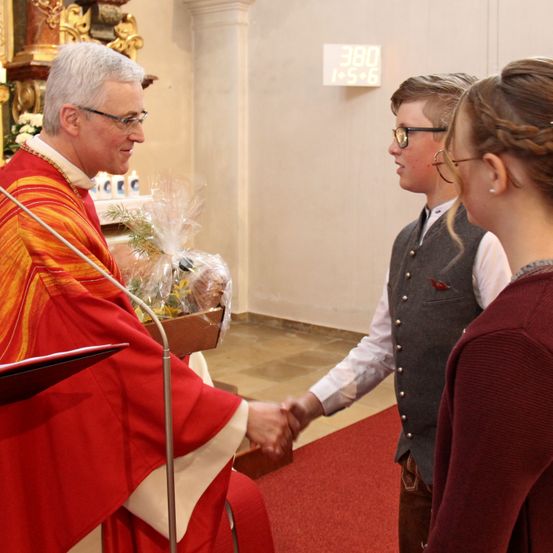 Ein Priester in einer roten Robe schüttelt einem jungen Jungen und einem Mädchen die Hand in einer Kirche.