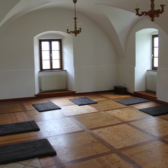 Ein leerer Raum mit Holzboden, mehreren Yoga-Matten und Bogenfenstern. Eine Kronleuchter hängt von der Decke.