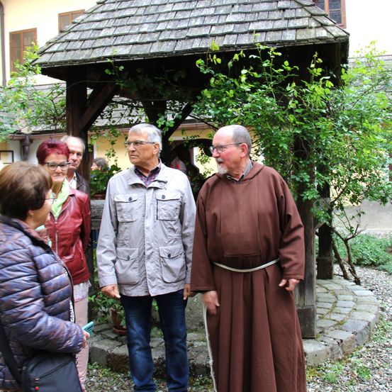 Eine Gruppe von Menschen steht in der Nähe eines Pavillons, mit zwei Männern vorne. Ein Mann trägt eine braune Robe und einen weißen Gürtel, während der andere eine Brille und eine Jacke trägt. Hinter ihnen stehen mehrere Menschen, darunter eine Frau in einer braunen Lederjacke.