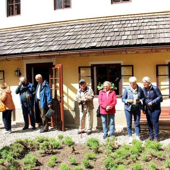 Eine Gruppe älterer Menschen steht vor einem Haus, einige machen Fotos und andere stehen um einen Garten herum.