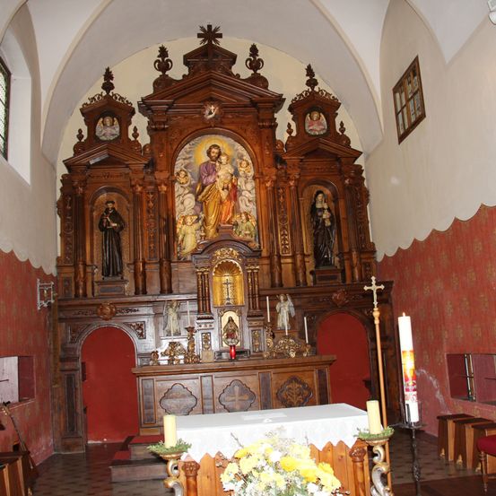 Der Altar einer Kirche zeigt ein großes zentrales Gemälde, das Jesus mit Kindern darstellt. Umgeben ist er von Statuen und kleineren Gemälden. Der Altar ist reich verziert mit kunstvollen Schnitzereien und goldenen Akzenten. Davor steht ein Tisch, der mit einem weißen Tuch bedeckt ist, und eine Blumenvase.