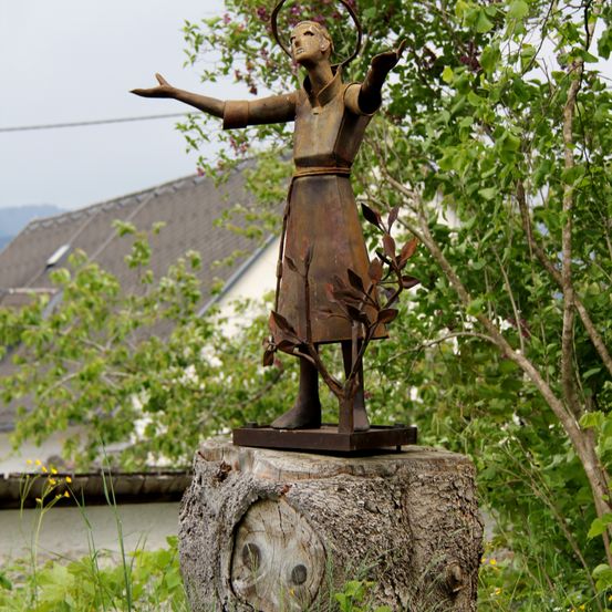 Eine Bronzestatue einer Frau steht mit erhobenen Armen auf einem Baumstumpf. Sie hat einen Heiligenschein über dem Kopf und ist von Bäumen und Pflanzen umgeben.