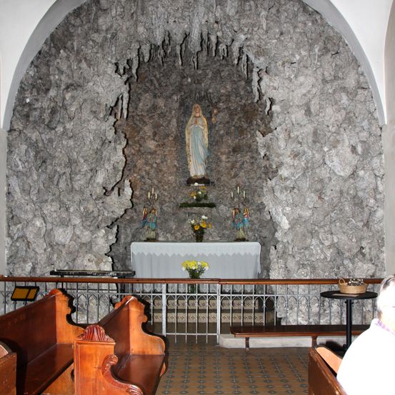 Eine Kapelle mit einer Statue der Jungfrau Maria hinter einer Felswand. Der Altar ist mit Kerzen, Blumen und Statuen geschmückt. Bänke und ein Geländer sind vorhanden.