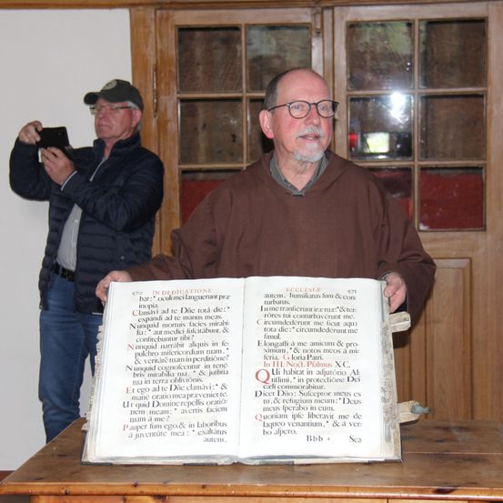 Ein älterer Mann hält ein offenes Buch mit lateinischem Text in einem Raum, während ein anderer Mann ein Foto macht.