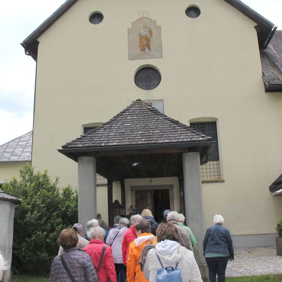 Eine Gruppe von Menschen steht vor einem Gebäude mit einem gefliesten Dach und einem hölzernen Eingang. Das Gebäude hat ein Bild eines Mannes an der Wand.
