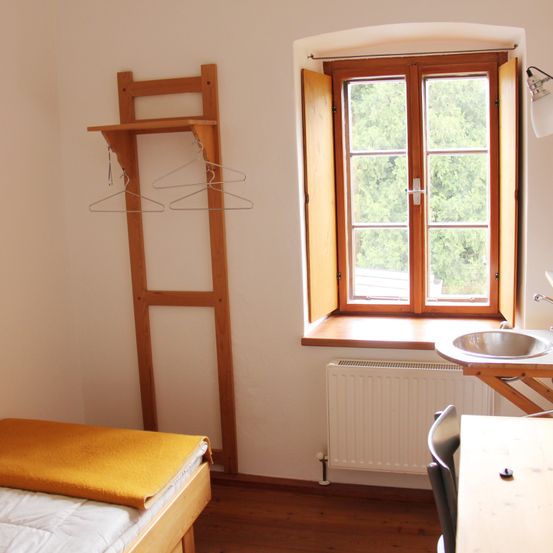 Ein kleines Schlafzimmer mit einem Einzelbett, einem Holzschrank und einem offenen Fenster mit Holzläden. Unter dem Fenster befindet sich ein Heizkörper, ein Schreibtisch mit Stuhl und ein Waschbecken auf einem Holzregal.