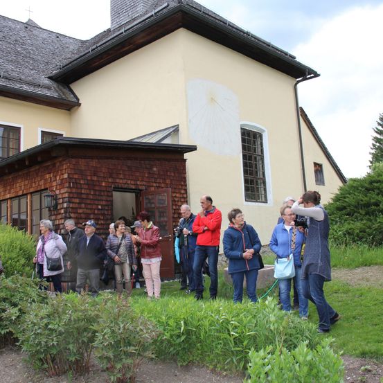 Eine Gruppe von Menschen steht vor einem Gebäude, einige halten Kameras und machen Fotos. Das Gebäude hat eine Sonnenuhr an der Seite und ein Fenster mit einem weißen Rahmen.
