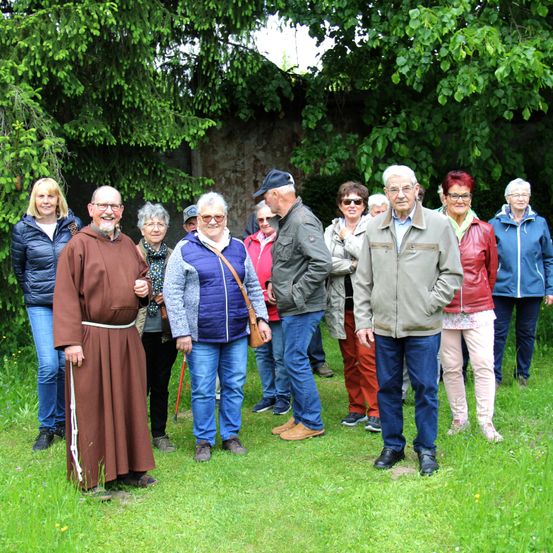 Eine Gruppe von Menschen in unterschiedlicher Kleidung steht in einem Park, mit einem Ordensbruder in einem braunen Gewand vorne. Bäume und Grünpflanzen sind hinter ihnen.