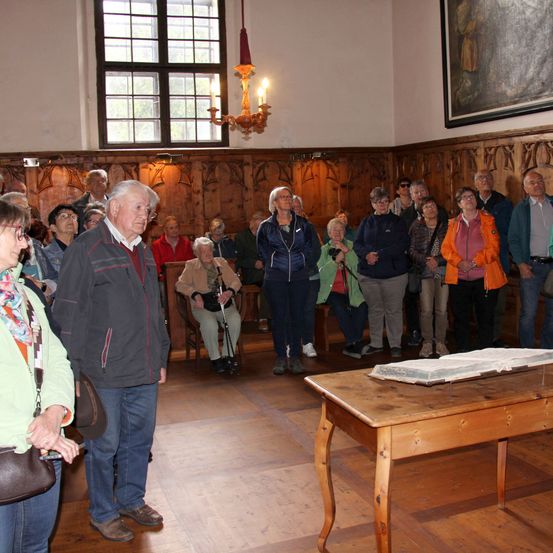 In einem Raum mit Holzwänden versammeln sich eine Gruppe von Menschen um einen Tisch. Ein älterer Mann steht vorne, und ein Buch liegt auf dem Tisch. Eine Laterne hängt von der Decke, und ein Gemälde schmückt die Wand.