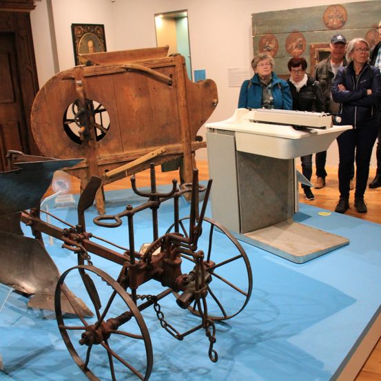 Ein alter Pflug und ein hölzerner Wagen werden auf einer blauen Plattform ausgestellt. Dahinter stehen Menschen und schauen zu. Der Hintergrund besteht aus einer weißen Wand mit einer Holztür.