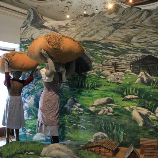 Zwei Puppen in traditioneller Kleidung tragen große Säcke auf ihren Schultern in einer Berglandschaft. Ein detailliertes Gemälde zeigt eine ländliche Szene mit Bergen, einem Holzhaus und verstreuten Felsen.