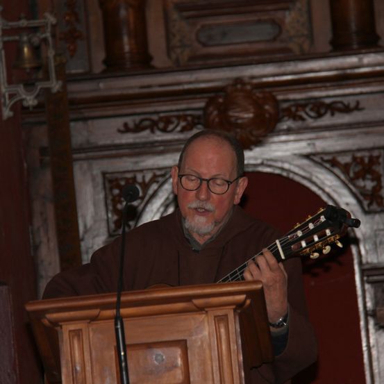 Ein älterer Mann in einer braunen Robe spielt eine Gitarre an einem Podium mit Mikrofon in einer Kirche.