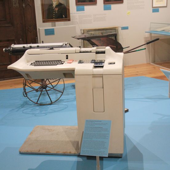 Eine Schreibmaschine ist in einem Museum ausgestellt, mit einer Holztür und einem Porträt im Hintergrund. Ein blauer Teppich liegt unter der Schreibmaschine.