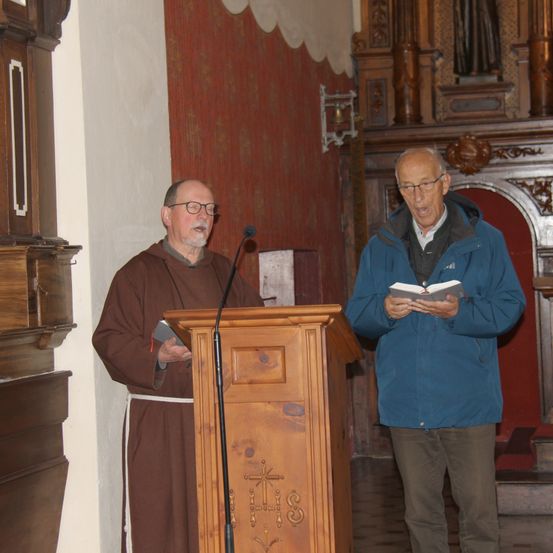 Zwei Männer stehen in einer Kirche. Einer liest aus einem Buch, während der andere aus einem Buch am Podium liest.