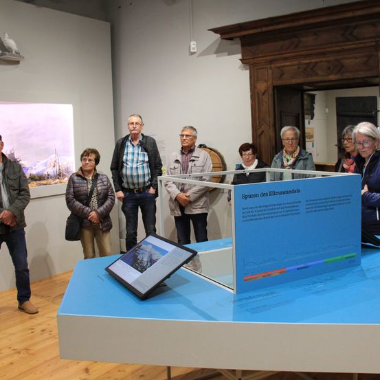 Eine Gruppe von Menschen steht um einen Ausstellungstisch mit einem Bildschirm und einer Glasbox herum. Sie scheinen eine Ausstellung in einem Museum zu betrachten.
