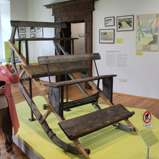Ein alter hölzerner Schlitten wird in einem Museum ausgestellt. Eine Person steht daneben und hält eine Kamera. An der Wand hinter dem Schlitten hängen mehrere gerahmte Bilder.