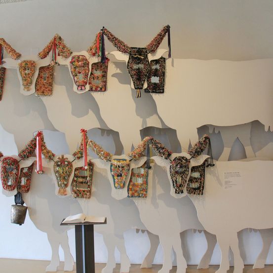 An einer Wand sind mehrere weiße Kuhskulpturen zu sehen, jede mit farbenfrohen, komplizierten Designs verziert. Die Kühe haben verschiedene Dekorationen, darunter Glocken und Bänder. Unter den Skulpturen steht ein offenes Buch auf einem Podest.