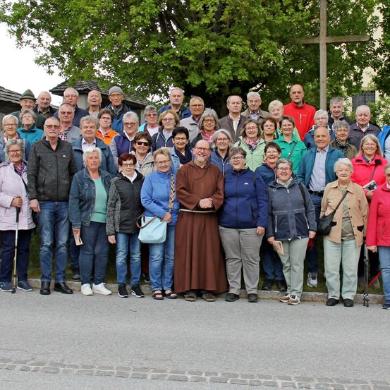 Eine große Gruppe von Menschen posiert für ein Foto auf der Straße, einige mit Brillen und Mützen. Ein Mann in einem braunen Gewand steht in der Mitte. Bäume und ein Kreuz stehen hinter ihnen.