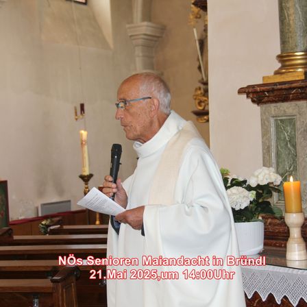 Ein älterer Priester in einer weißen Robe und mit einer Brille hält ein Mikrofon und liest aus einem Stück Papier in einer Kirche. Hinter ihm befinden sich brennende Kerzen, Blumen und ein hölzerner Altar. Das Datum und die Uhrzeit der Veranstaltung sind auf dem Bild zu sehen.